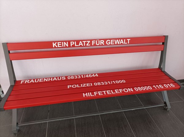 Abgebildet ist eine Stadtbank in roter Farbe. Auf ihr sind Telefonnummern in weißer Farbe augemalt z. B. die des Frauenhauses in Memmingen 08331 / 4644.
