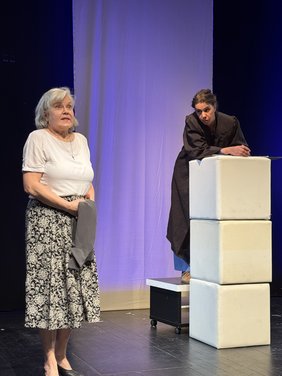 Bühnenszene: Eine Frau vorne redet, eine andere weiter hinten hört zu. Violetter Hintergrund.